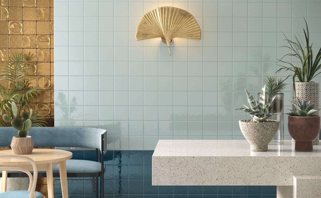 Bottega d'Arte TerreLucide Oro 3 espace bistrot avec des carreaux de faïence Bottega d'Arte TerreLucide Oro bleu clair et doré, applique murale en forme d'éventail doré, table en terrazzo blanc avec des plantes en pot, chaise avec coussins bleus et table ronde en bois.