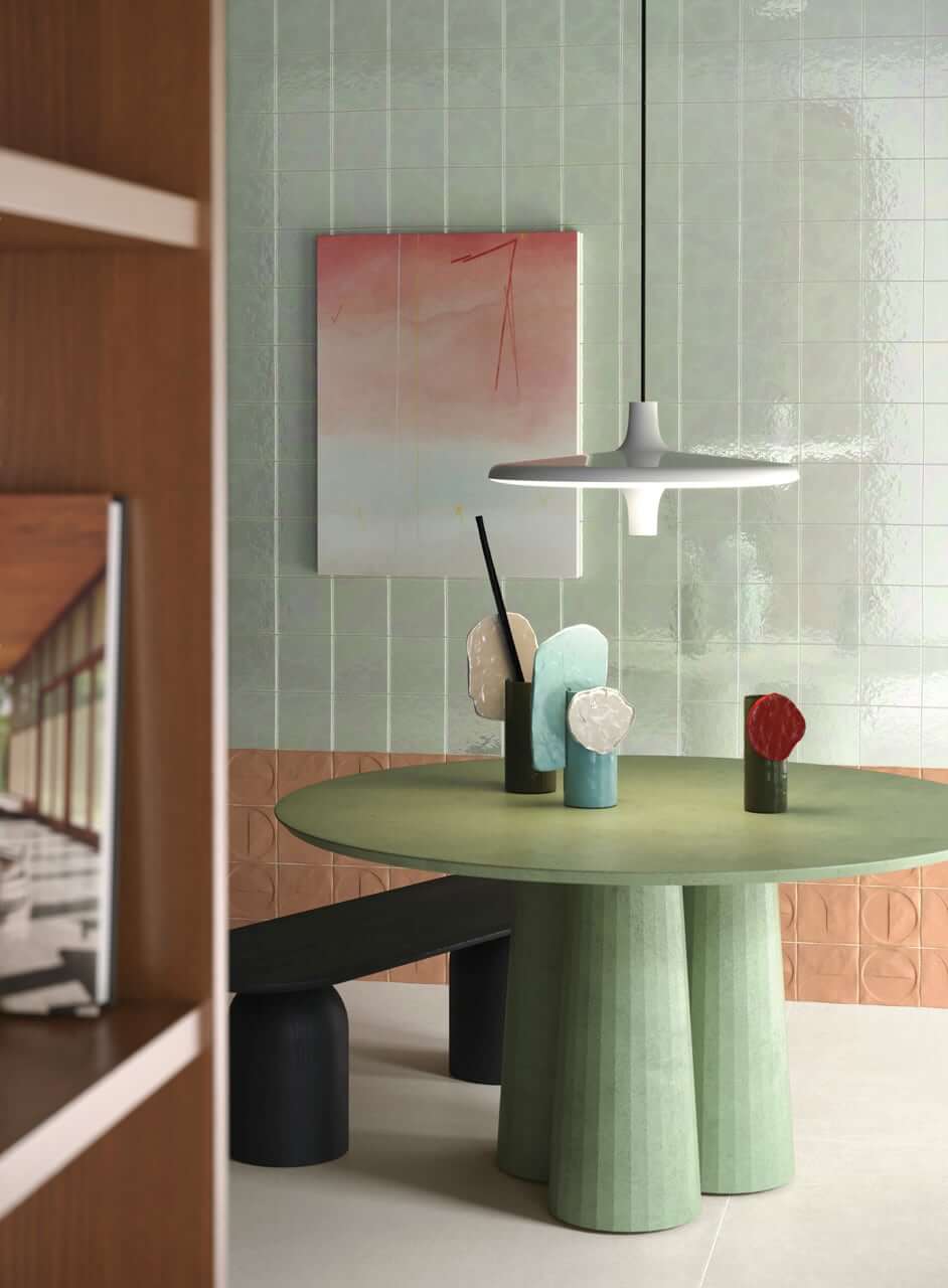 Bottega d'Arte TerreLustrate 3 espace de café-bar avec une table ronde vert pâle, carreaux de faïence Bottega d'Arte TerreLustrate vert pâle et orange clair sur le mur, vases en céramique artistique sur la table, lampe suspendue blanche au-dessus, tableau abstrait sur le mur.